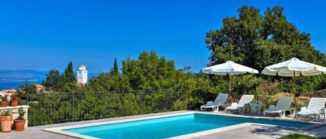 A rectangular swimming pool with white lounge chairs and umbrellas, surrounded by a paved patio and a metal fence.