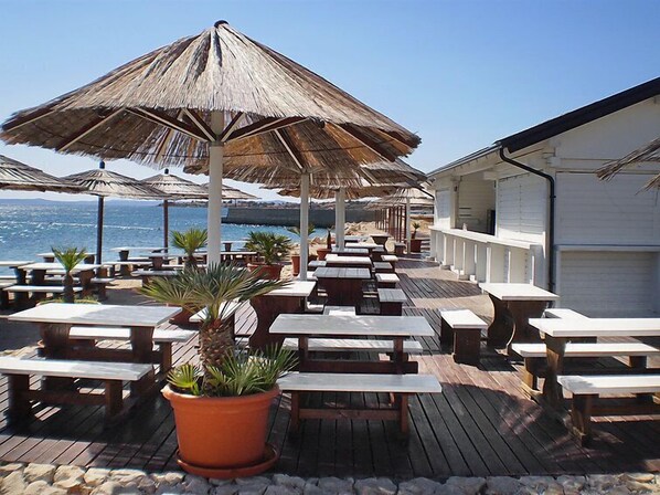 A beachside restaurant with wooden tables and thatched umbrellas, overlooking the sea.