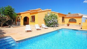 Zona de piscina con una piscina azul cristalina, sillas de descanso blancas y un edificio amarillo con tejado de baldosas.