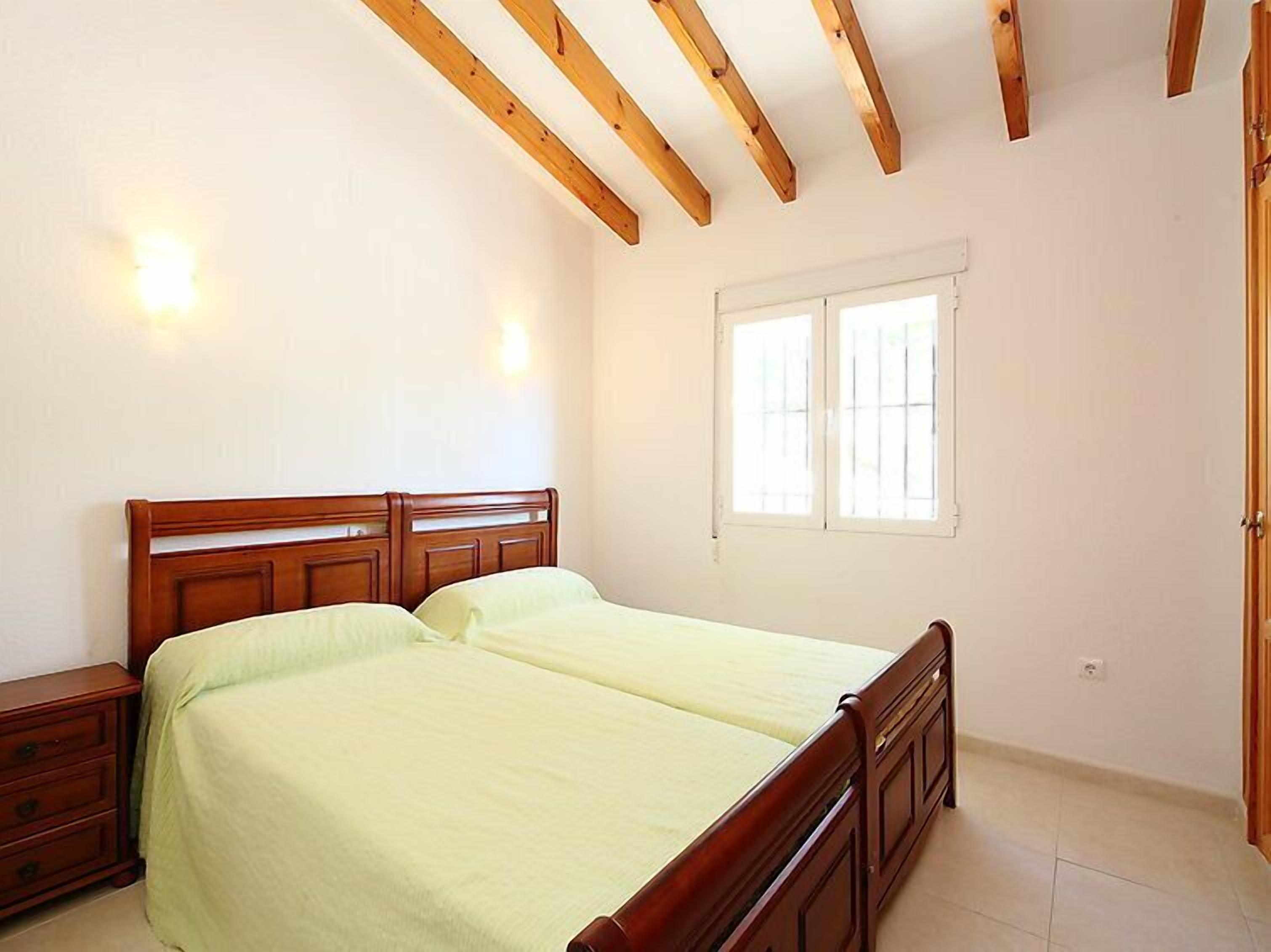 A bedroom with a wooden bed frame, two bedside tables, a window with bars, and a door.