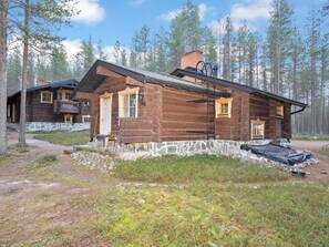 Une cabane rustique en rondins de bois, avec des fondations en pierre, une cheminée et une véranda dotée d’un banc.