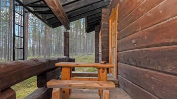 A wooden cabin with a bench and a table, surrounded by a wooden deck and a forest backdrop.