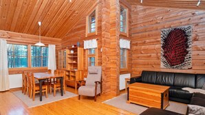 A log cabin interior with a dining area, a living room with a black leather sofa, and a large window with a view of trees.
