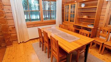 Een houten chalet van binnen met een eettafel, stoelen, een kast en uitzicht op het bos door grote ramen.