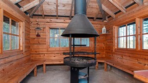 A log cabin interior with a central wood stove, wooden walls, and large windows.