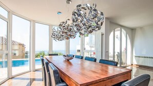 A spacious dining area with a wooden table and black leather chairs, large windows offering a view of a swimming pool and a building across the water. - Vacation home Gaudí by Interhome (Lloret de Mar)