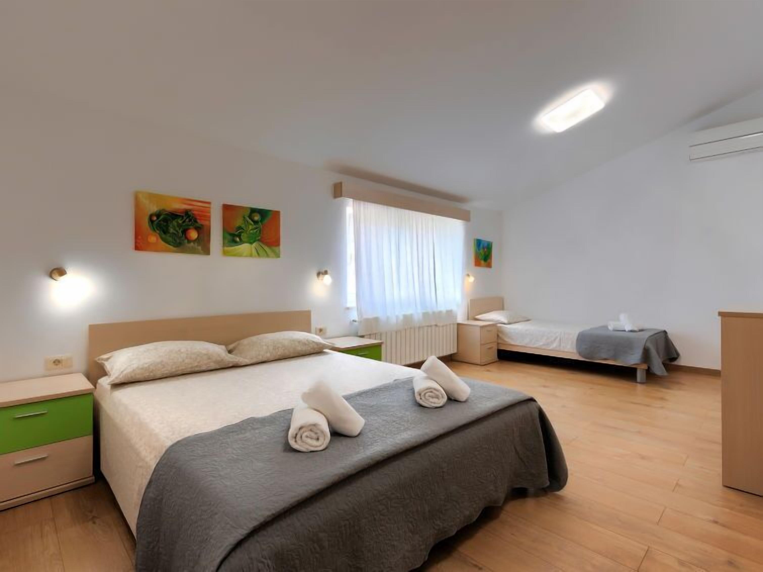 Une chambre d’hôtel avec deux lits, une table de chevet verte, une commode en bois et une fenêtre avec des rideaux blancs.