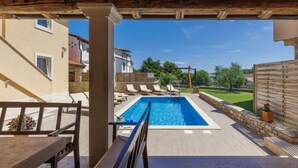 A covered outdoor swimming pool with lounge chairs and a table, surrounded by a stone wall and a wooden fence. - Licardo by Interhome (Štinjan)