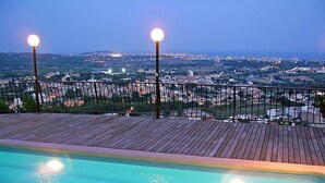 A pool with a wooden deck and railing, overlooking a cityscape at dusk. - Mas Ambros by Interhome (Calonge)