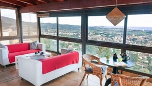 A spacious room with a large window offering a city view, a white sofa with red cushions, wicker chairs, and a small table with a kettle and cups. - Mas Ambros by Interhome (Calonge)