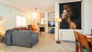 A living room with a sofa, a dining table with chairs, a fireplace, a television, and a large painting. - Bonalba Golf, Urb. Los Naranjos by Interhome (San Juan / Muchamiel)
