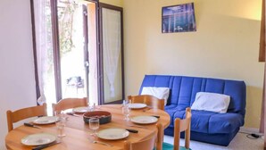 Ein Essbereich mit einem runden Holztisch, gedeckt für vier Personen, umgeben von Holzstühlen mit türkisfarbenen Kissen. Eine blaue Couch mit weißen Kissen lehnt an der Wand, und darüber ist ein Poster zu sehen.