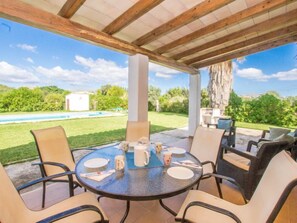 A covered outdoor patio with a round table and chairs, a pool in the background, and a large tree.