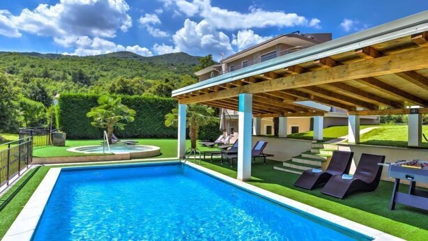 A swimming pool with a wooden deck and lounge chairs, surrounded by lush greenery and a mountain backdrop. - Vacation home Nicole by Interhome (Opatija/Pobri)