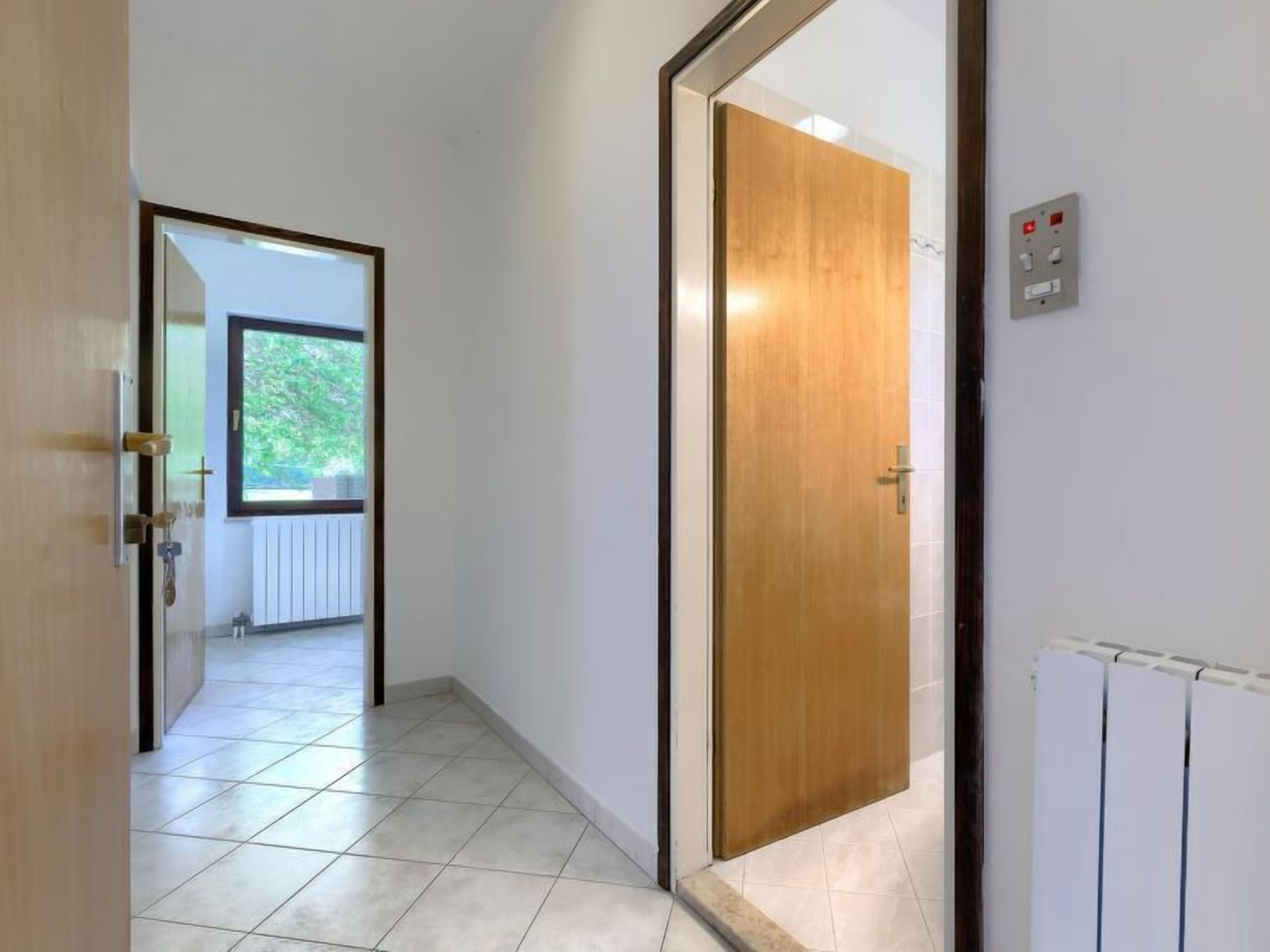 Un pasillo con una puerta de madera, un radiador y una ventana con vista a la vegetación.