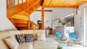 Una sala de estar moderna con una escalera de madera, un sofá con cojines estampados, sillas de mimbre y una mesa de centro de madera con una planta.