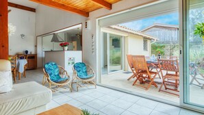 Un amplio salón con un gran ventanal corredera que da a un patio exterior. En el interior, hay un sofá, dos sillones de mimbre con cojines azules y una mesa de centro de madera.