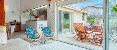 A spacious living area with a large sliding glass door leading to an outdoor patio. Inside, there is a sofa, two wicker chairs with blue cushions, and a wooden coffee table.