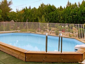 A rectangular swimming pool with a wooden deck and metal railings, surrounded by a white fence and trees.