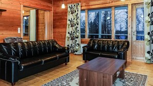 L’intérieur d’une cabane en rondins est aménagé avec deux canapés en cuir noir, une table basse en bois et de grandes fenêtres qui offrent une vue sur les arbres.