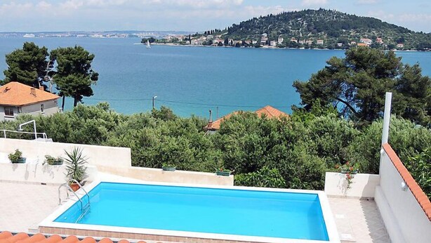 Une piscine sur le toit offrant une vue sur un paysage côtier, entourée de verdure et de bâtiments.
