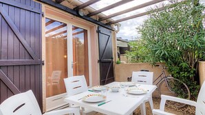 Um pátio externo com mesa posta para quatro pessoas, rodeado por cadeiras brancas, sob uma pérgula de madeira.