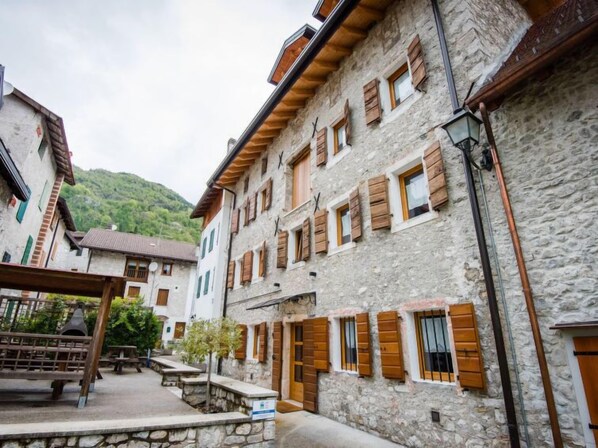 A stone building with wooden shutters and a balcony, a stone bench, and a small table in a courtyard.