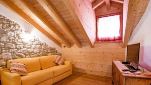 A cozy attic room with a wooden ceiling, a stone wall, a yellow sofa, and a wooden cabinet with a computer. - Albergo Diffuso - Cjasa Ustin by Interhome (Barcis Lake)