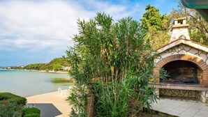A stone-built outdoor kitchen area with a brick oven, surrounded by lush greenery and a view of a calm body of water. - Apartment Mare 8 by Interhome (Rab/Kampor)