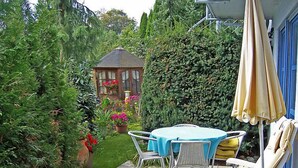 Una veranda con tavolo e sedie, immersa nel verde e con un piccolo gazebo sullo sfondo.