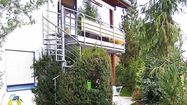 Un edificio residenziale con una scala visibile che conduce a un balcone, circondato da un giardino curato e ricco di piante e fiori.