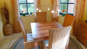 Zona pranzo con tavolo in legno e sedie in vimini, due finestre con tende, una lampada a sospensione e un vaso con piante secche.