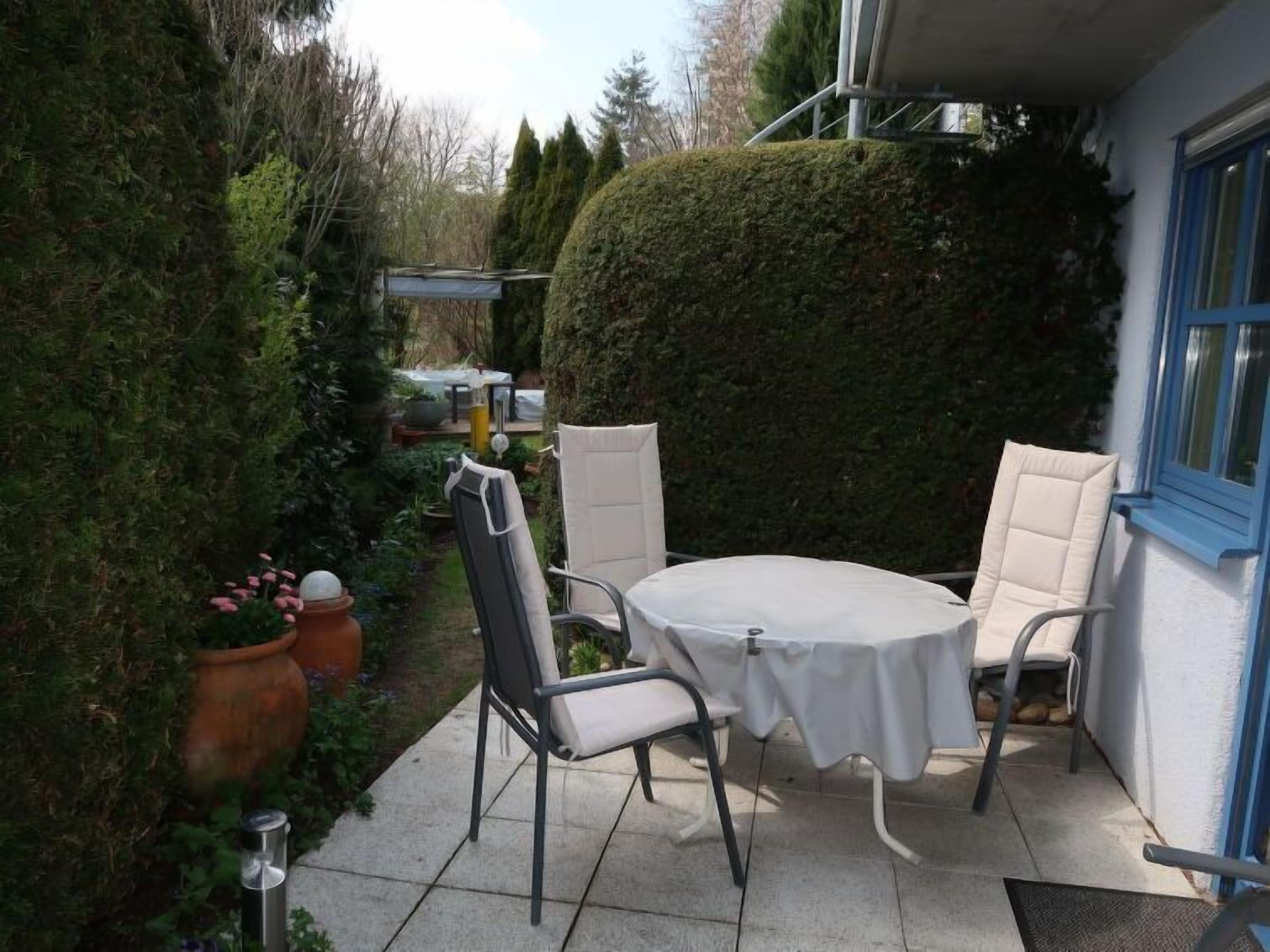 Een terras met een ronde tafel en twee stoelen, omringd door groen en een witte muur.