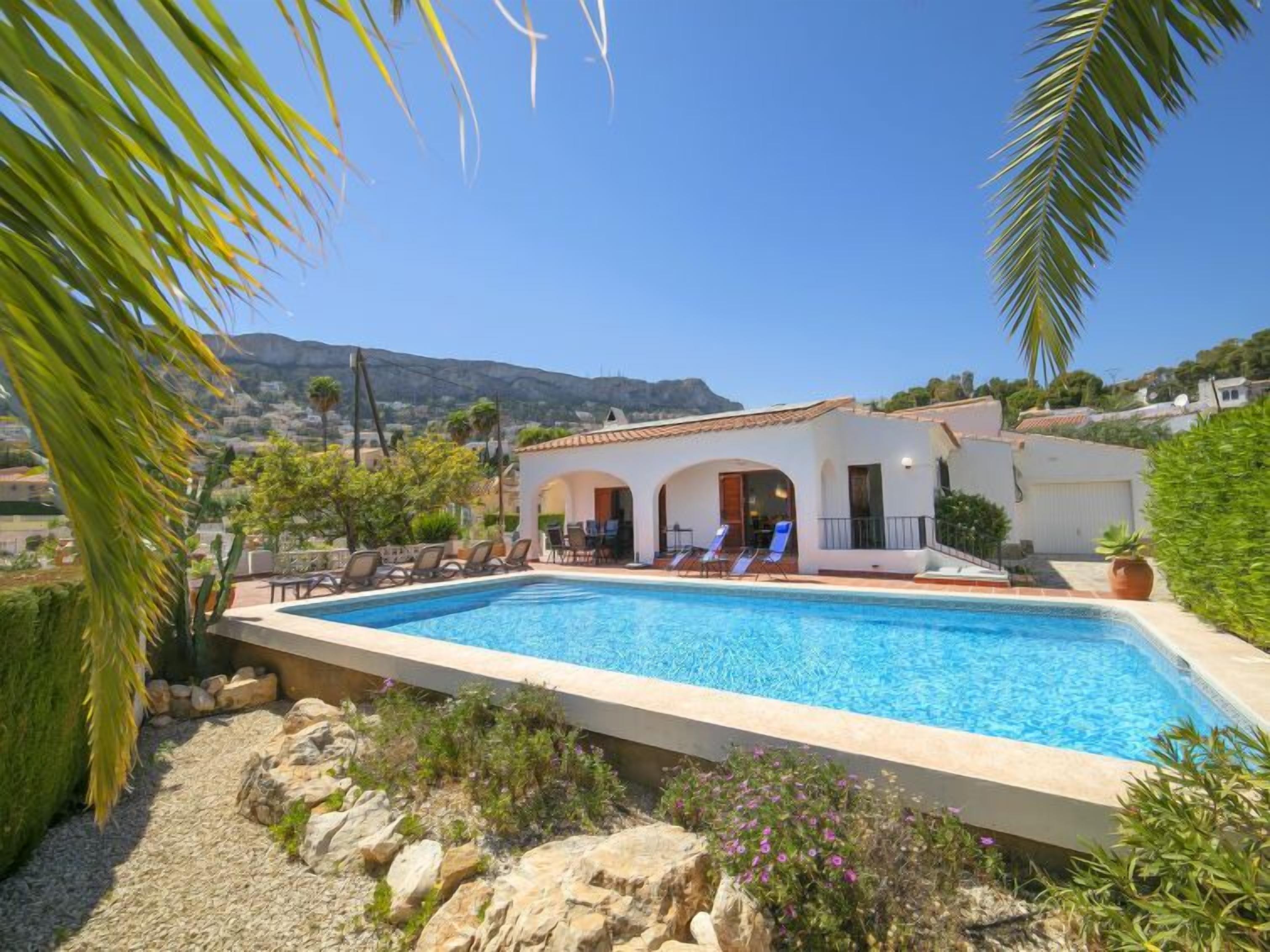 A poolside area with a clear view of a mountain in the background, a white building with a tiled roof, and a seating area with lounge chairs.