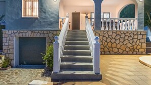 Una casa de dos plantas con fachada de piedra, una escalera central con balaustres blancos y un patio con baldosas.