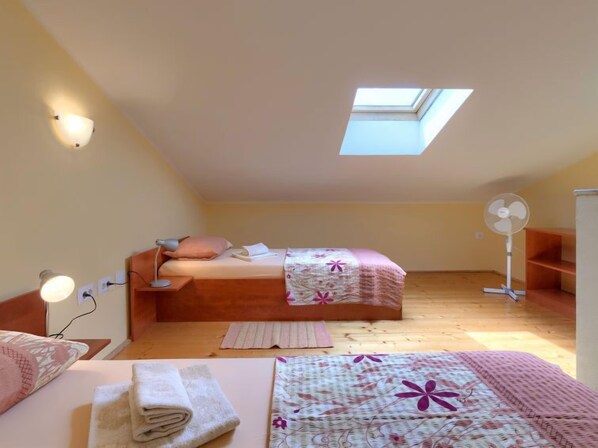 Ein kleiner, gemütlicher Raum mit einem Oberlicht, einem Bett mit floralem Überwurf, einem Nachttisch mit Lampe und einem Standventilator.