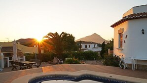 En swimmingpool med ett berg i bakgrunden, omgiven av utemöbler och en vit byggnad.
