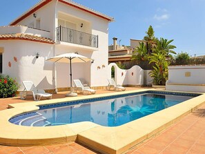 Una piscina con sillones de descanso y sombrillas, un edificio blanco con balcón y un patio con baldosas.