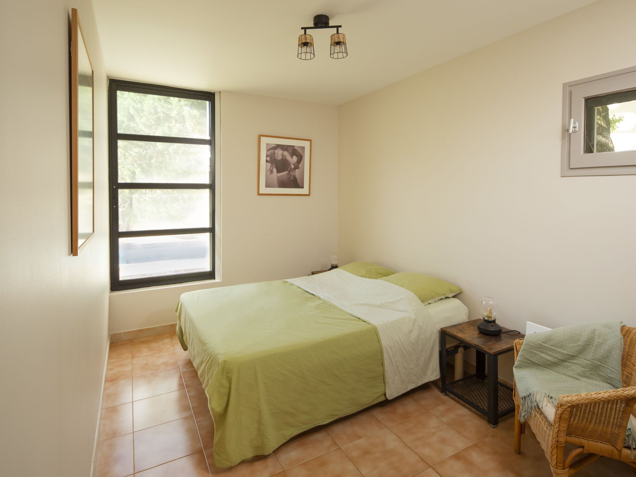 A bedroom with a bed, a small table, a wicker chair, a window, and a wall-mounted picture.