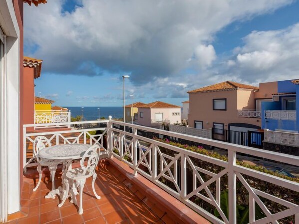 A balcony with a white metal railing, a round table with chairs, and a view of the sea and neighboring buildings.