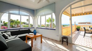 Quarto iluminado pelo sol com sofá, uma mesa pequena e uma cadeira. Grandes janelas oferecem vista para áreas verdes e uma varanda com mobiliário para exterior.