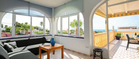 Habitación soleada con sofá, una mesita y una silla. Amplias ventanas ofrecen vistas a la vegetación y un balcón con mobiliario exterior.