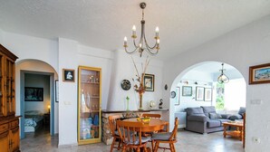 Uma sala de jantar ampla, com mesa e cadeiras de madeira, um lustre e vista através de um arco para uma sala de estar com sofá e mesa de centro.