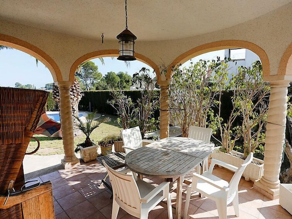 Un patio couvert avec une table ronde et des chaises blanches, entouré d’arcades et de plantes en pot.