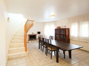 Un espace repas comprenant une longue table et des chaises, une cheminée, un escalier en bois et un meuble avec des portes en verre.