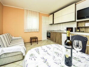 A modern living room with a sofa and a dining table set for two, a kitchen with wooden cabinets and a flat-screen TV, and a bottle of wine on the table.