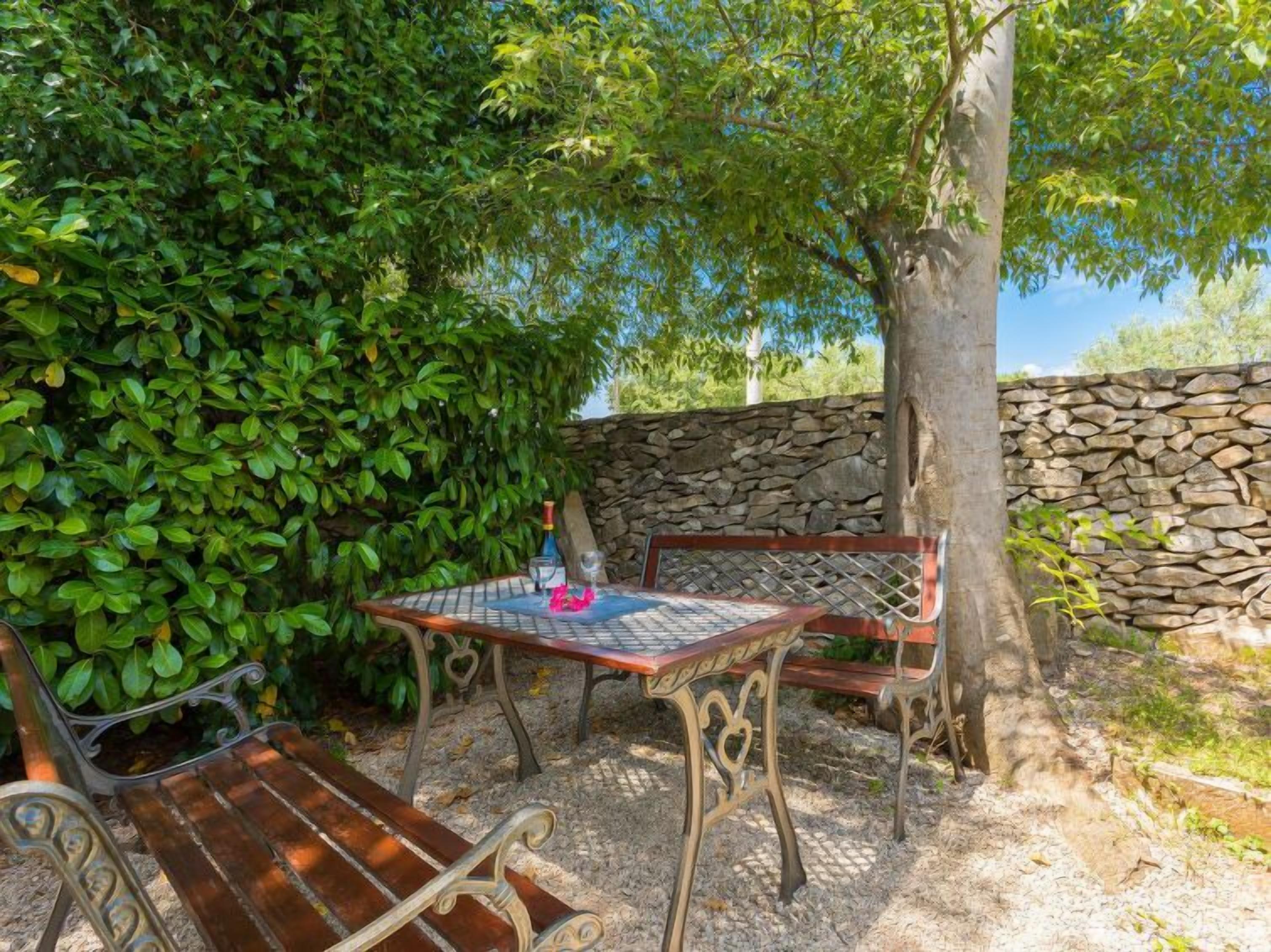 An outdoor seating area with a table and benches, surrounded by greenery and a stone wall.
