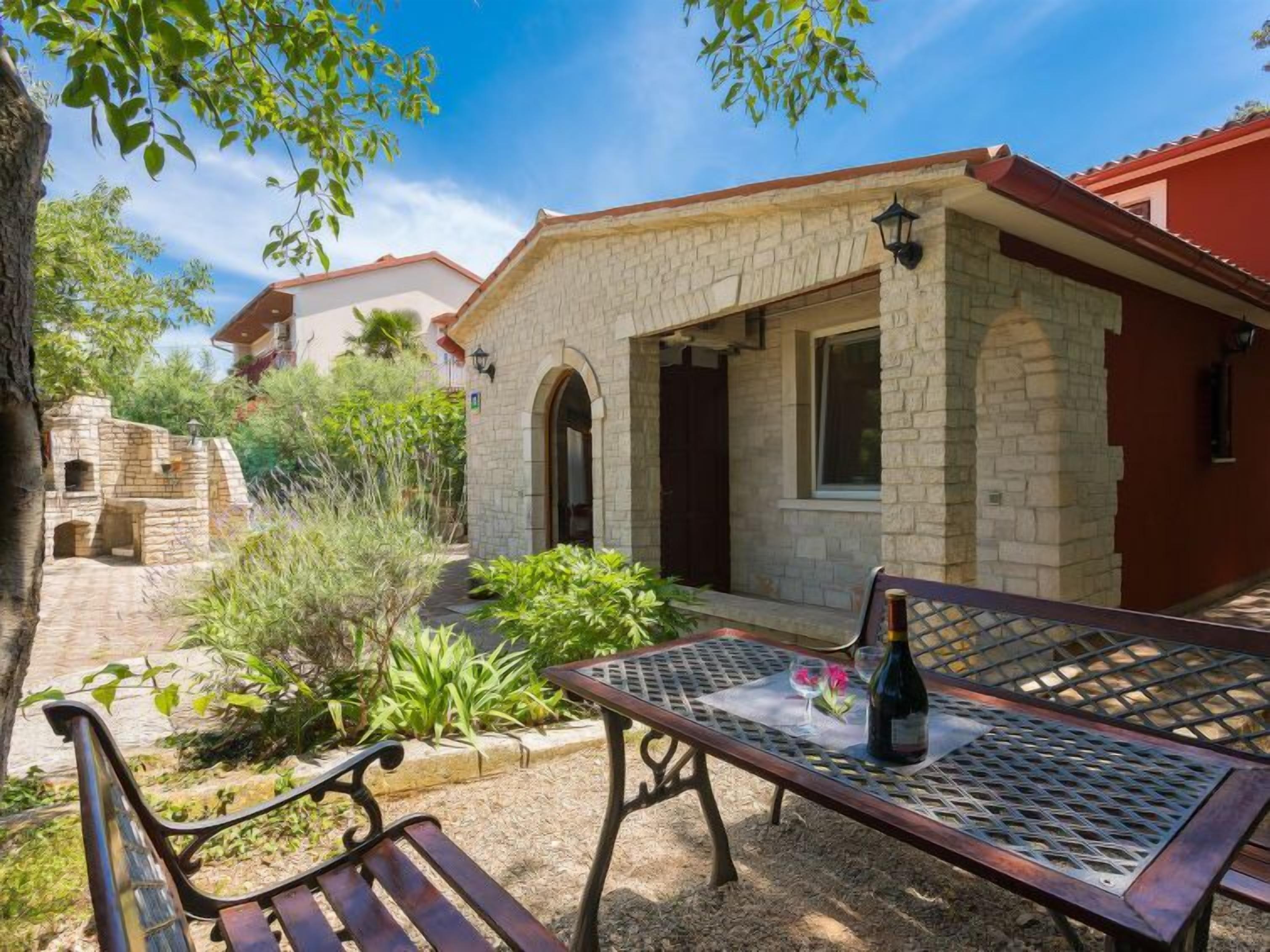 A stone building with a red roof, a paved patio with a metal table and bench, and a bottle of wine on the table.