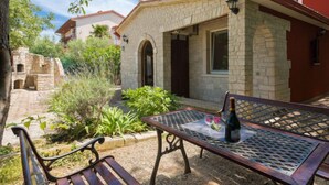 Un edificio de piedra con techo rojo, un patio empedrado con una mesa y banca de metal, y una botella de vino sobre la mesa.