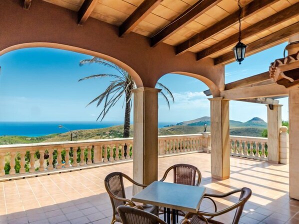 A covered outdoor seating area with a view of the sea and hills.